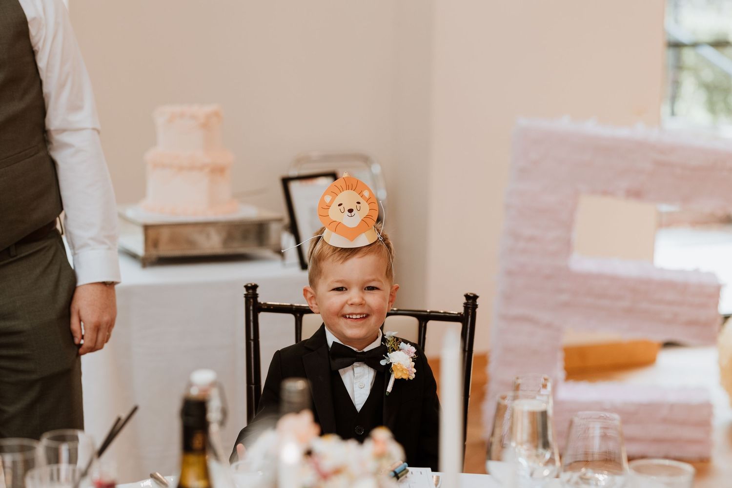A smiling young boy in a tuxedo with a lion hat sits at a wedding reception table, surrounded by elegant decor.