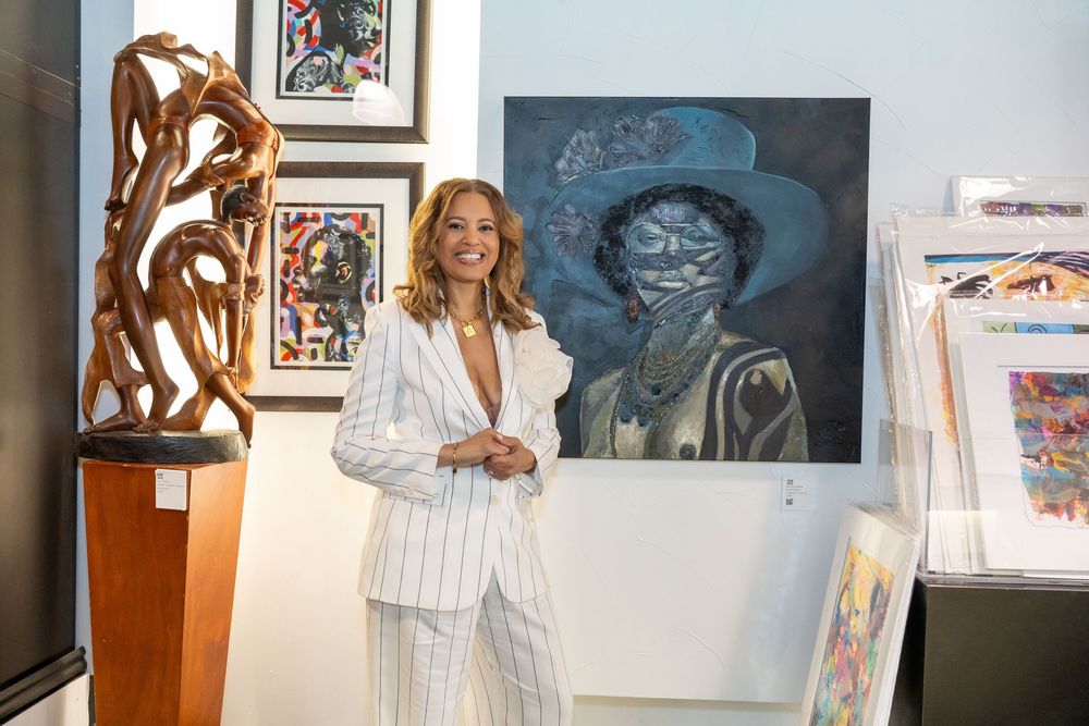 Smiling woman in white pinstripe suit standing among colorful artwork in an art gallery.