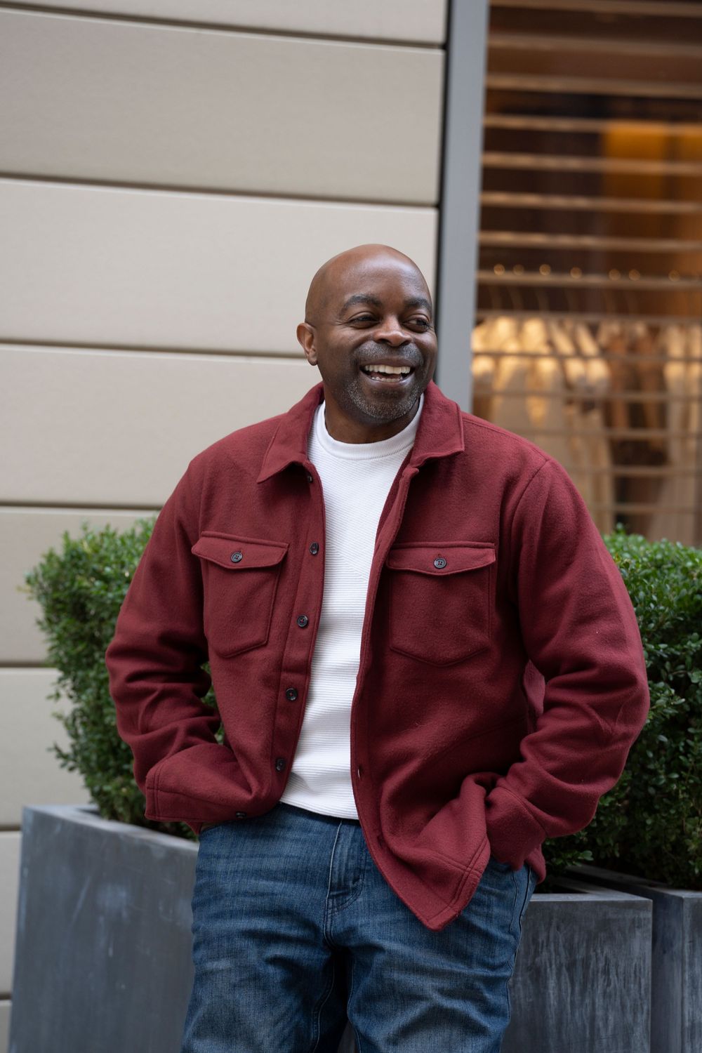 Happy bald man in red jacket and jeans smiling outdoors near green shrubs.