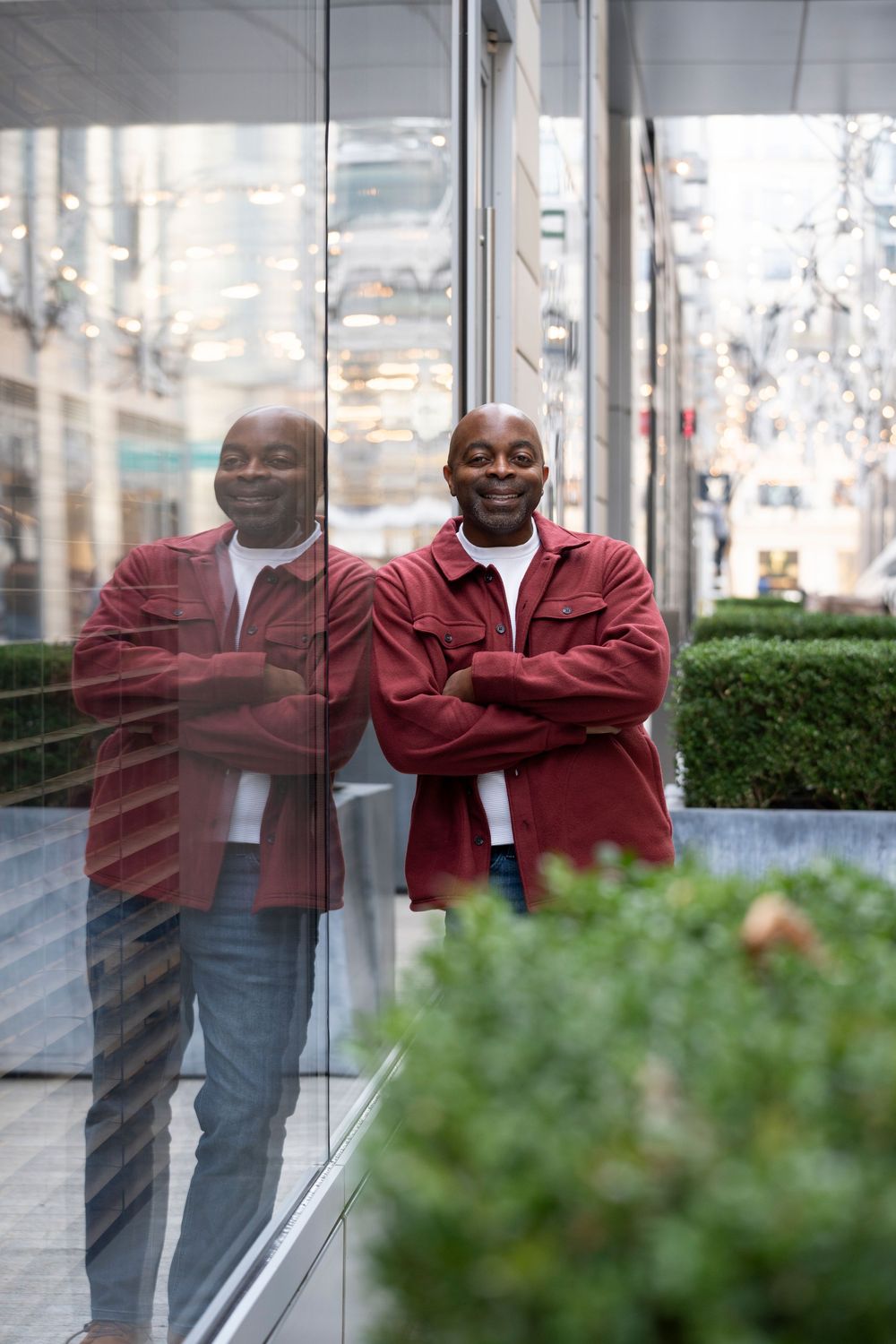 Smiling bald man in red jacket leaning against glass wall with reflection visible.