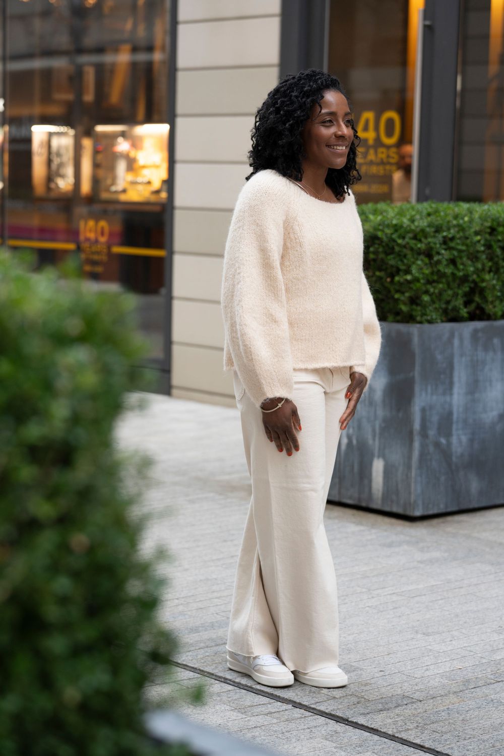 Woman in cream fuzzy sweater and wide-leg pants smiling on urban sidewalk.