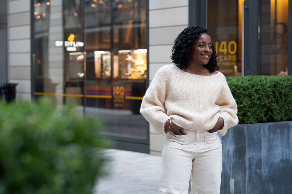 Smiling woman in cream sweater and white jeans standing outside a city storefront.