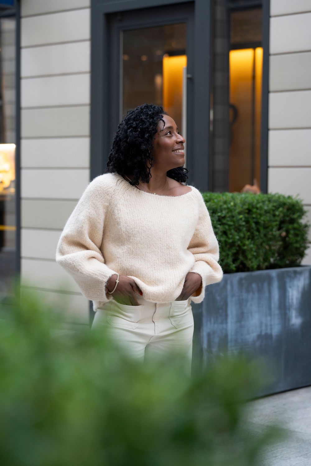 Stylish Black woman in cream sweater and white pants standing outside a modern building.