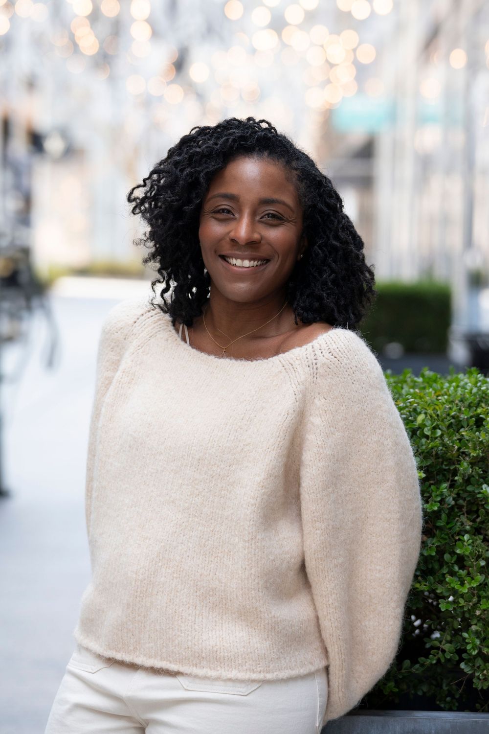 Smiling Black woman with curly hair wearing a cream knit sweater outdoors with bokeh lights.