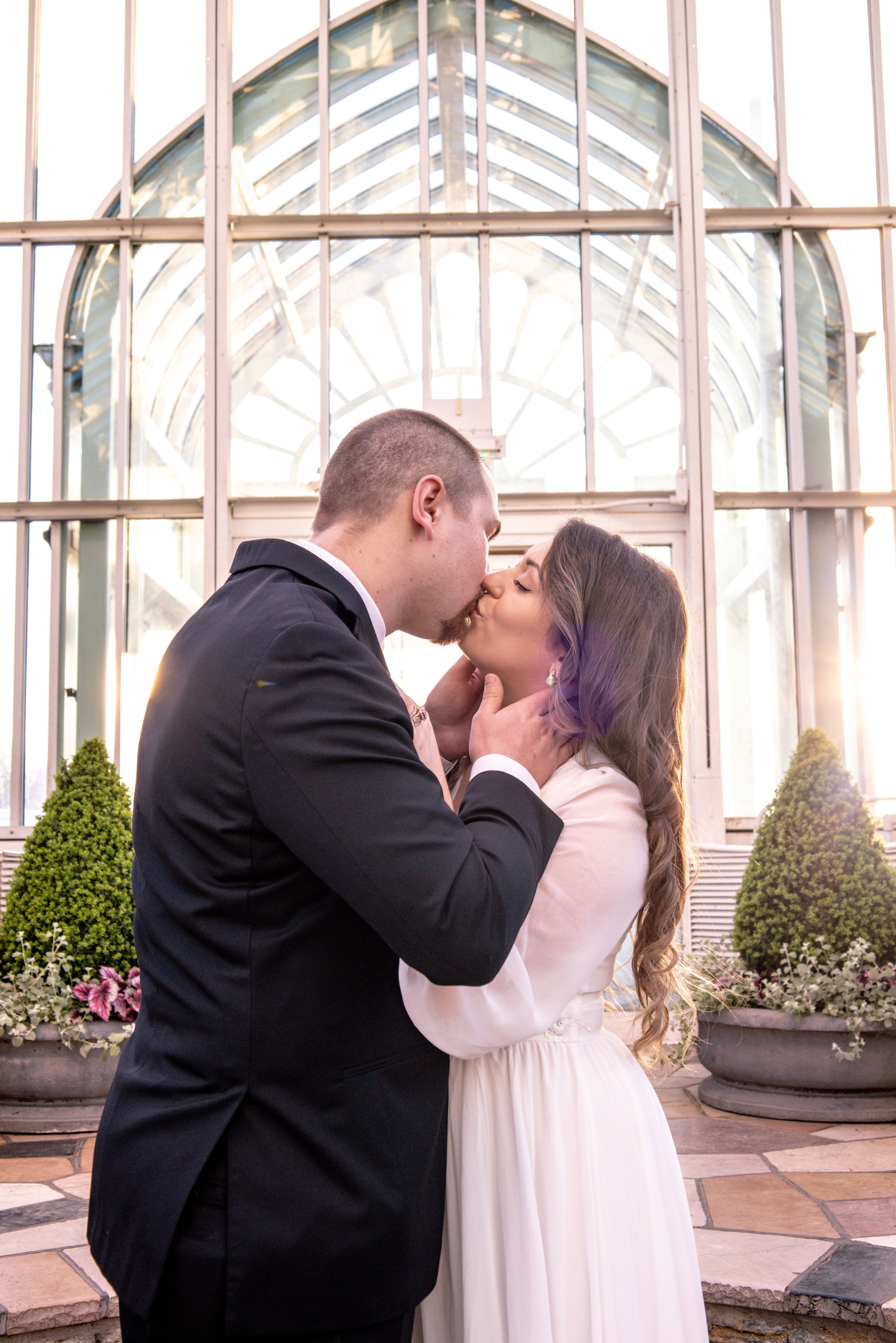 A couple shares a romantic kiss in front of a glass greenhouse, bride in white gown and groom in black suit.