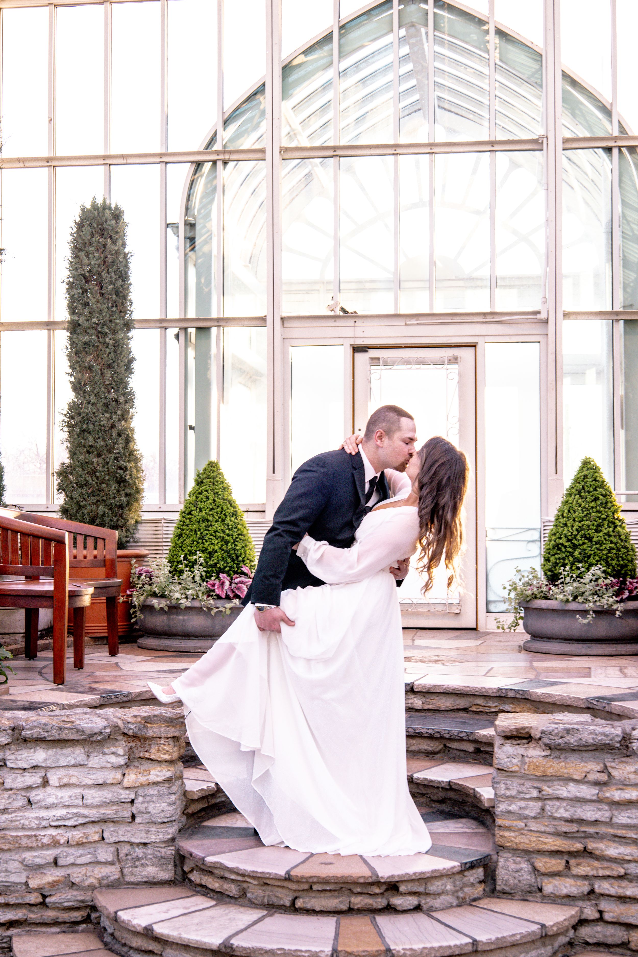 A bride and groom share a romantic dip kiss on stone steps in front of a glass greenhouse venue.