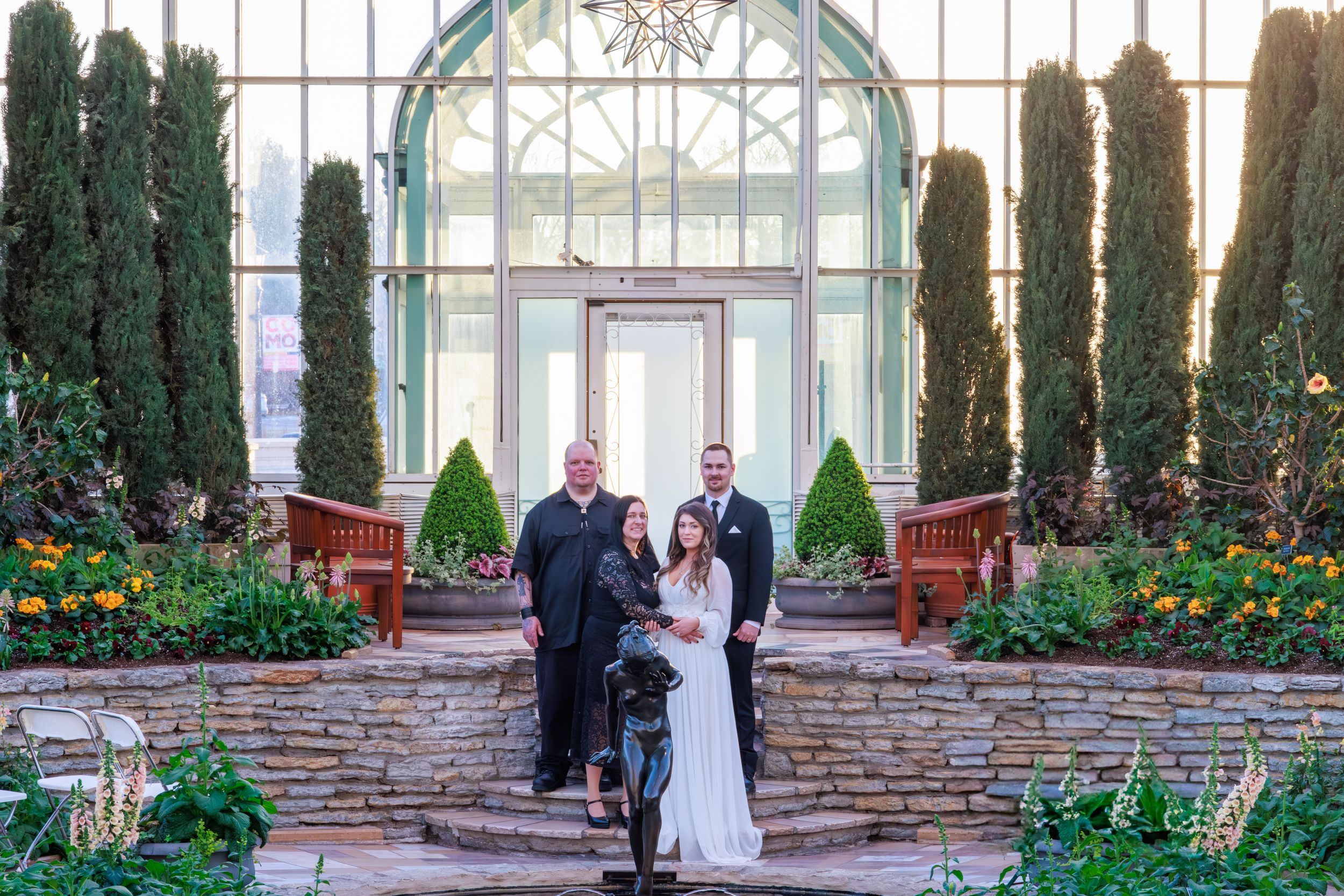 Wedding ceremony in a botanical garden greenhouse with couple, officiant, and guest on stone steps.