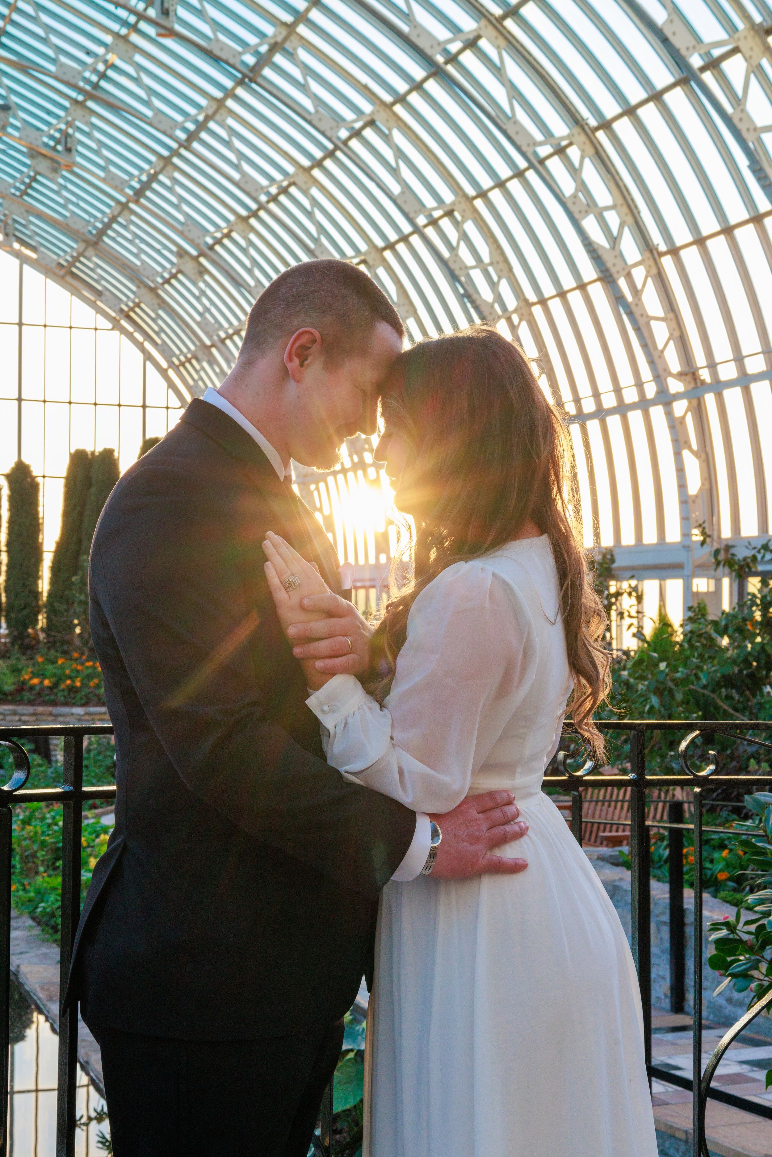 Bride and groom share a romantic moment in a sunlit glass greenhouse, golden light glowing behind them.