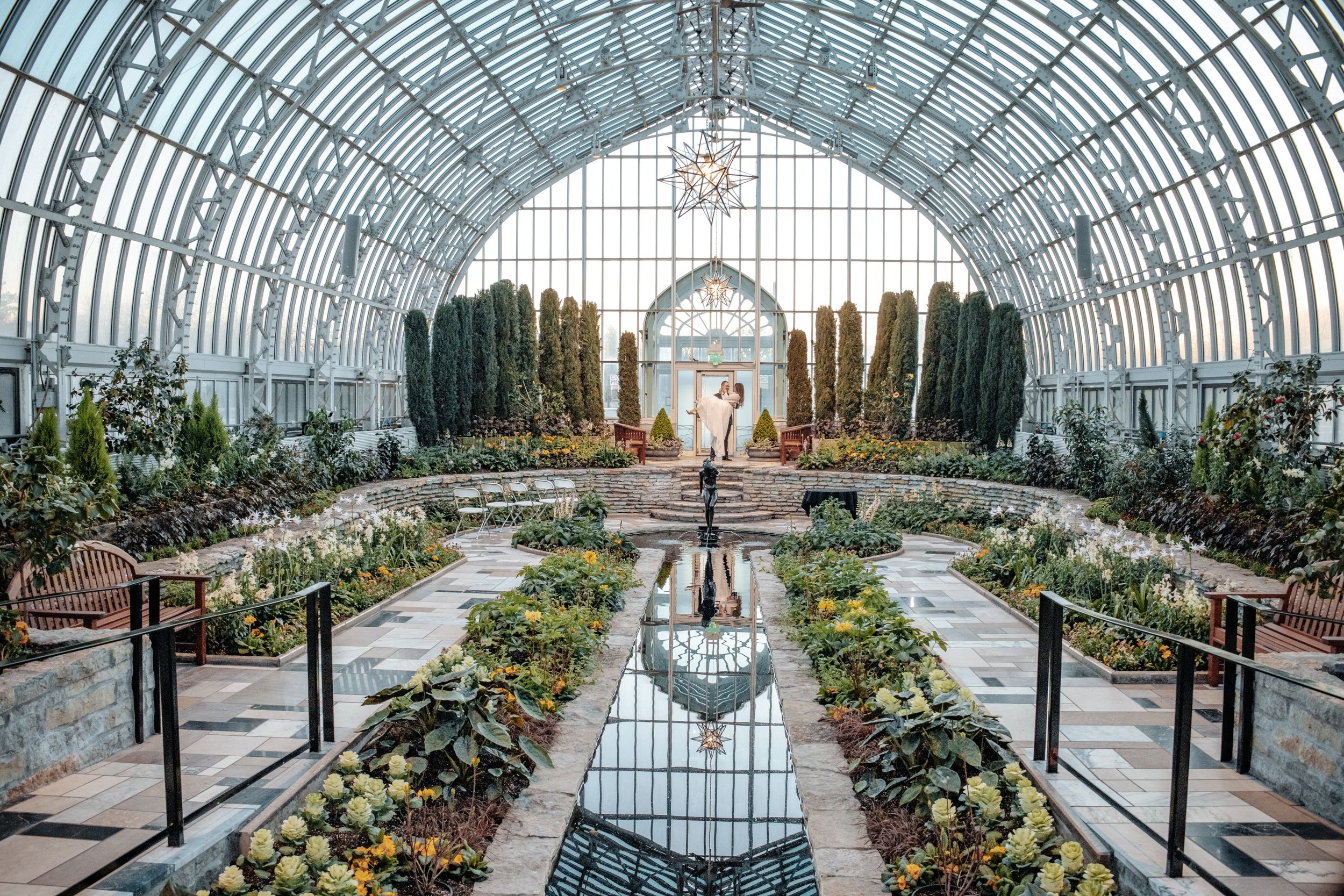 Couple stands in stunning greenhouse conservatory with arched glass ceiling, lush gardens, and elegant chandelier.