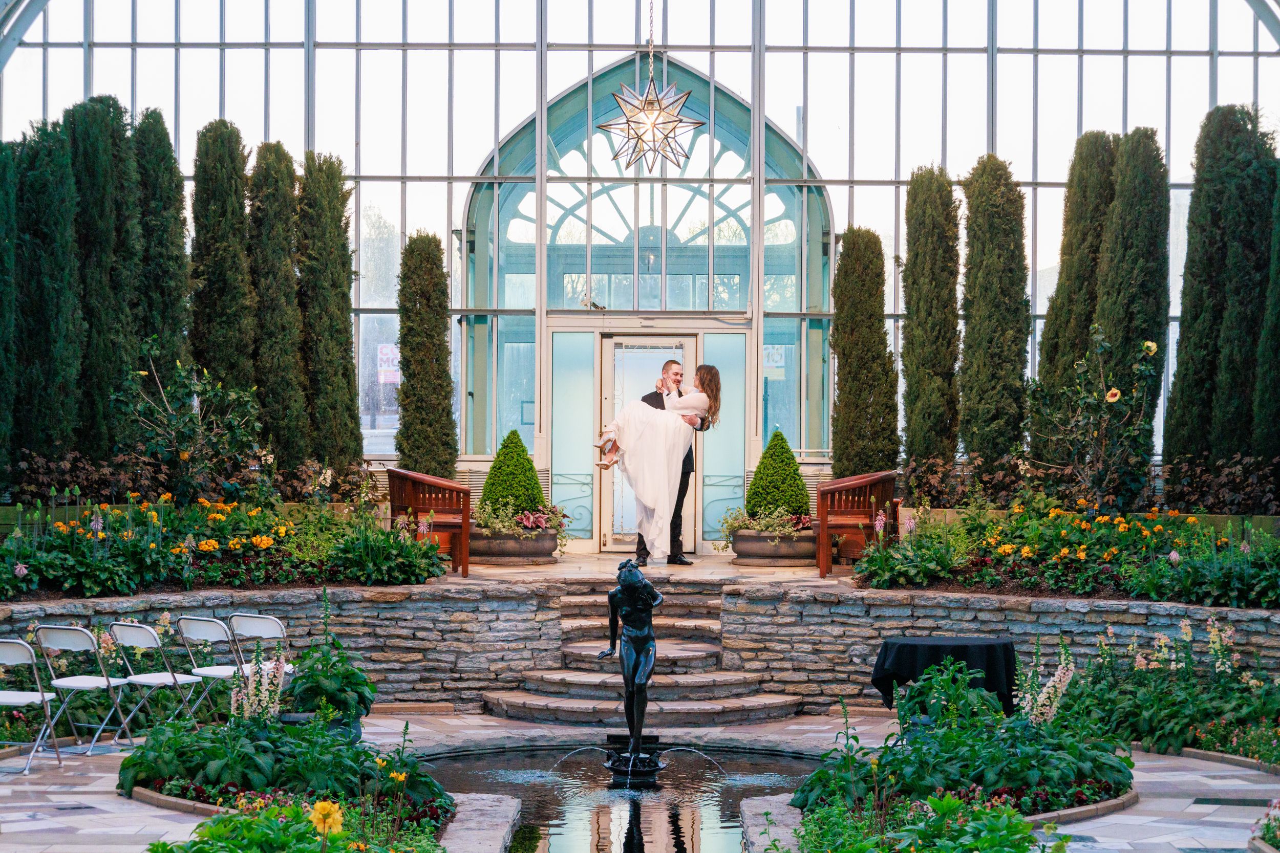 Couple embraces near fountain in elegant glass conservatory garden with cypress trees and colorful flowers.