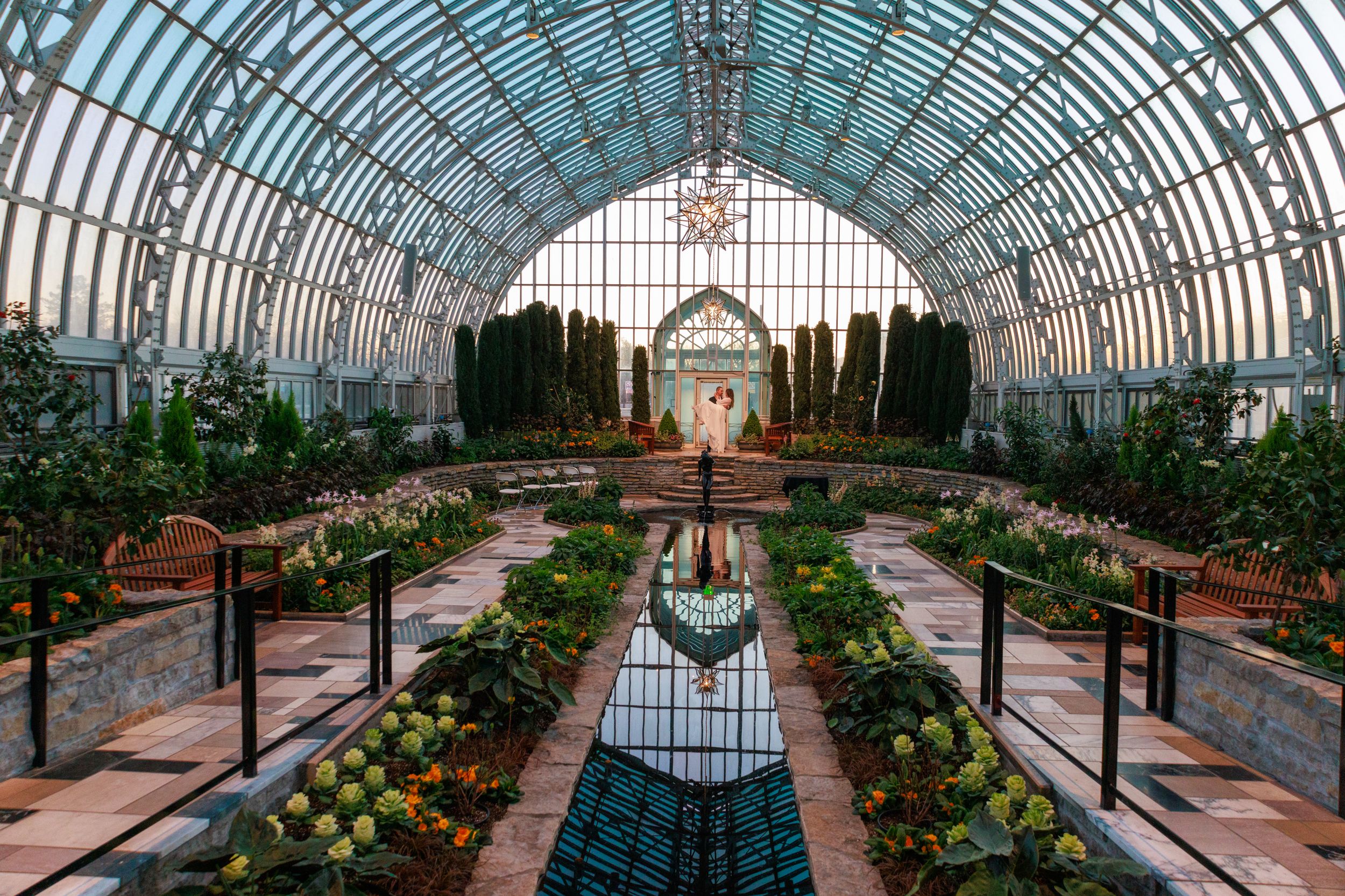 Stunning greenhouse conservatory with arched glass ceiling, lush gardens, reflecting pool, and ornate architecture in botanical garden.