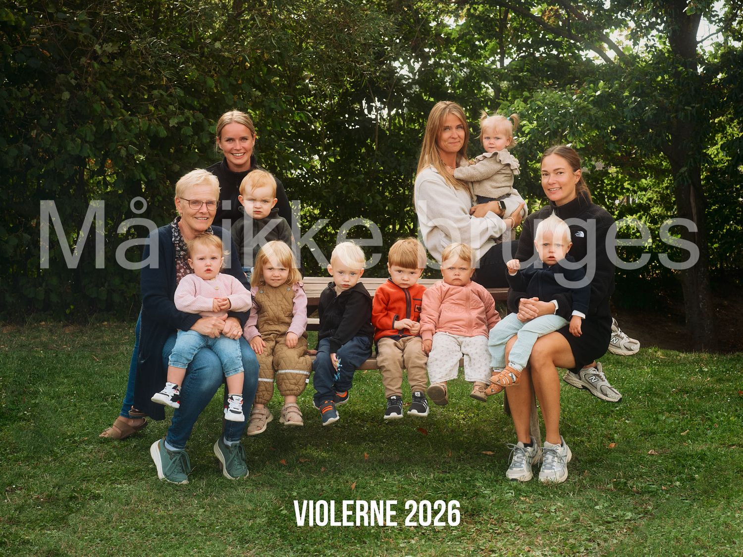 Family portrait of grandparents, parents, and young children posing outdoors on a bench in a green garden, labeled Violerne 2026.