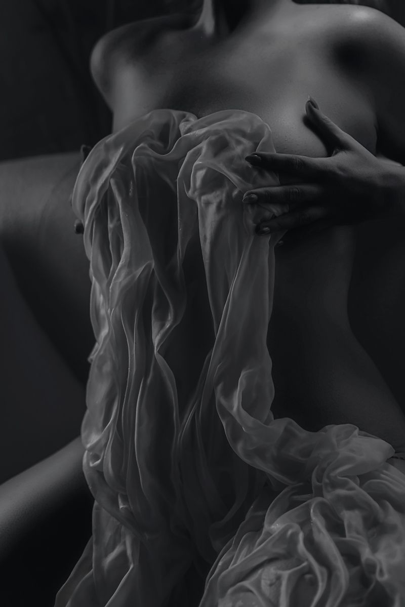 Black and white fine art photo of a woman draped in sheer flowing fabric, captured in dramatic chiaroscuro lighting.