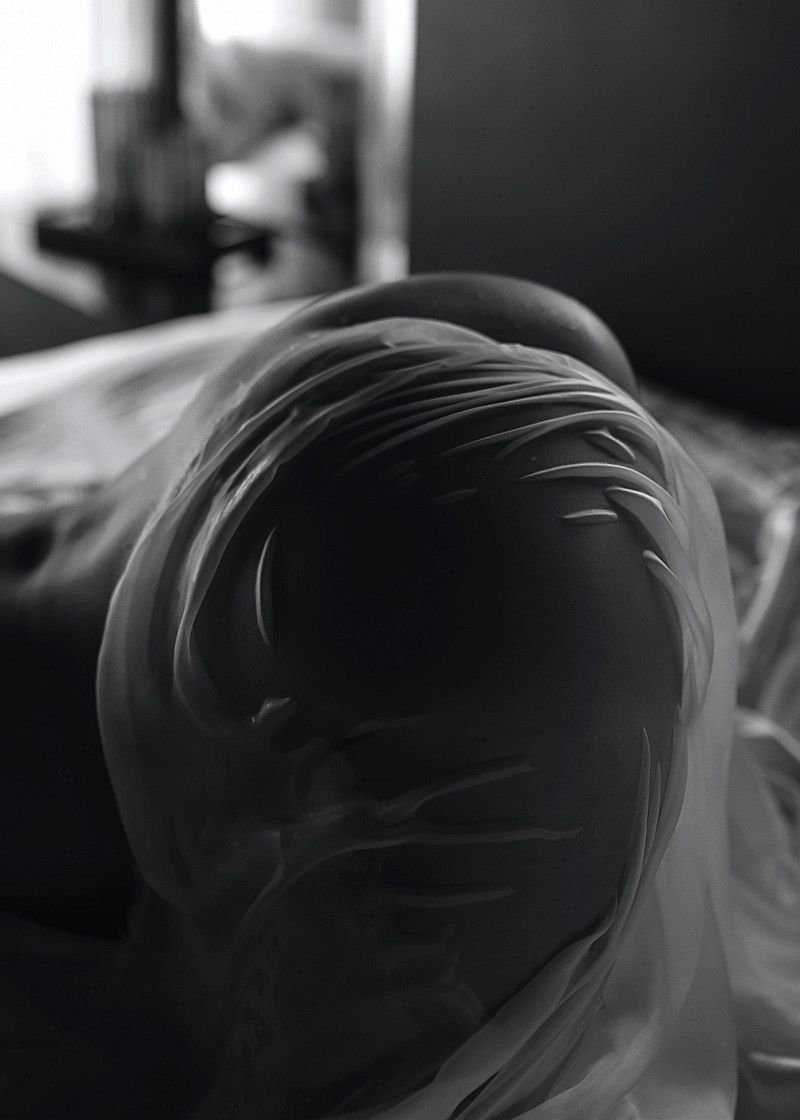 Black and white close-up photo of a human head wrapped in translucent plastic wrap, creating an eerie abstract effect.