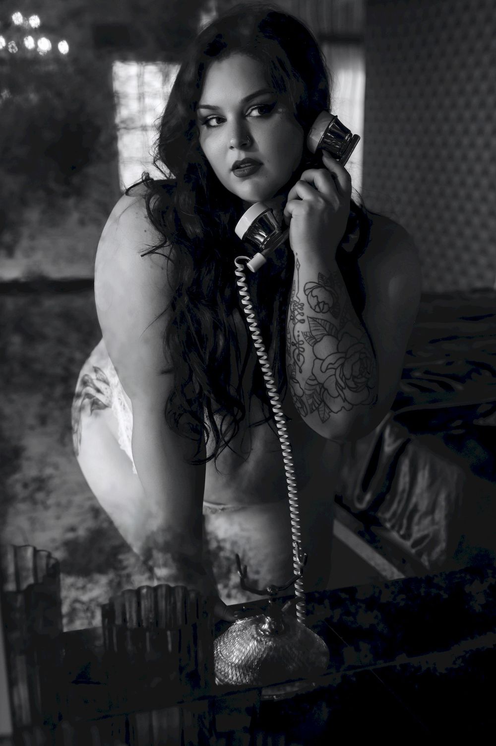 Black and white photo of a tattooed woman posing with a vintage rotary telephone in a moody, dimly lit setting.