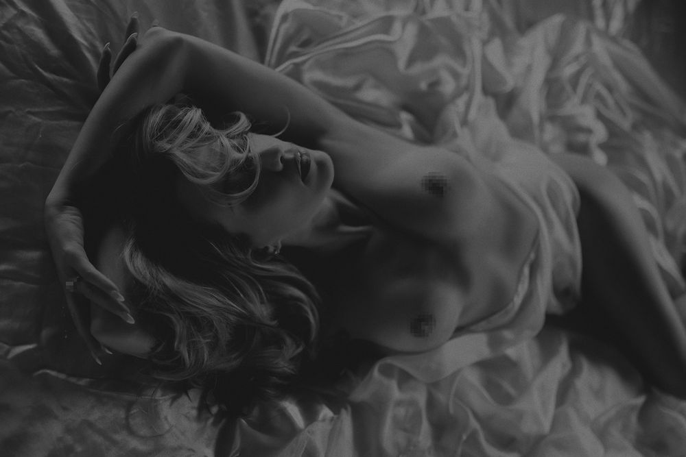 Black and white artistic nude photograph of a woman reclining gracefully on soft fabric bedding.