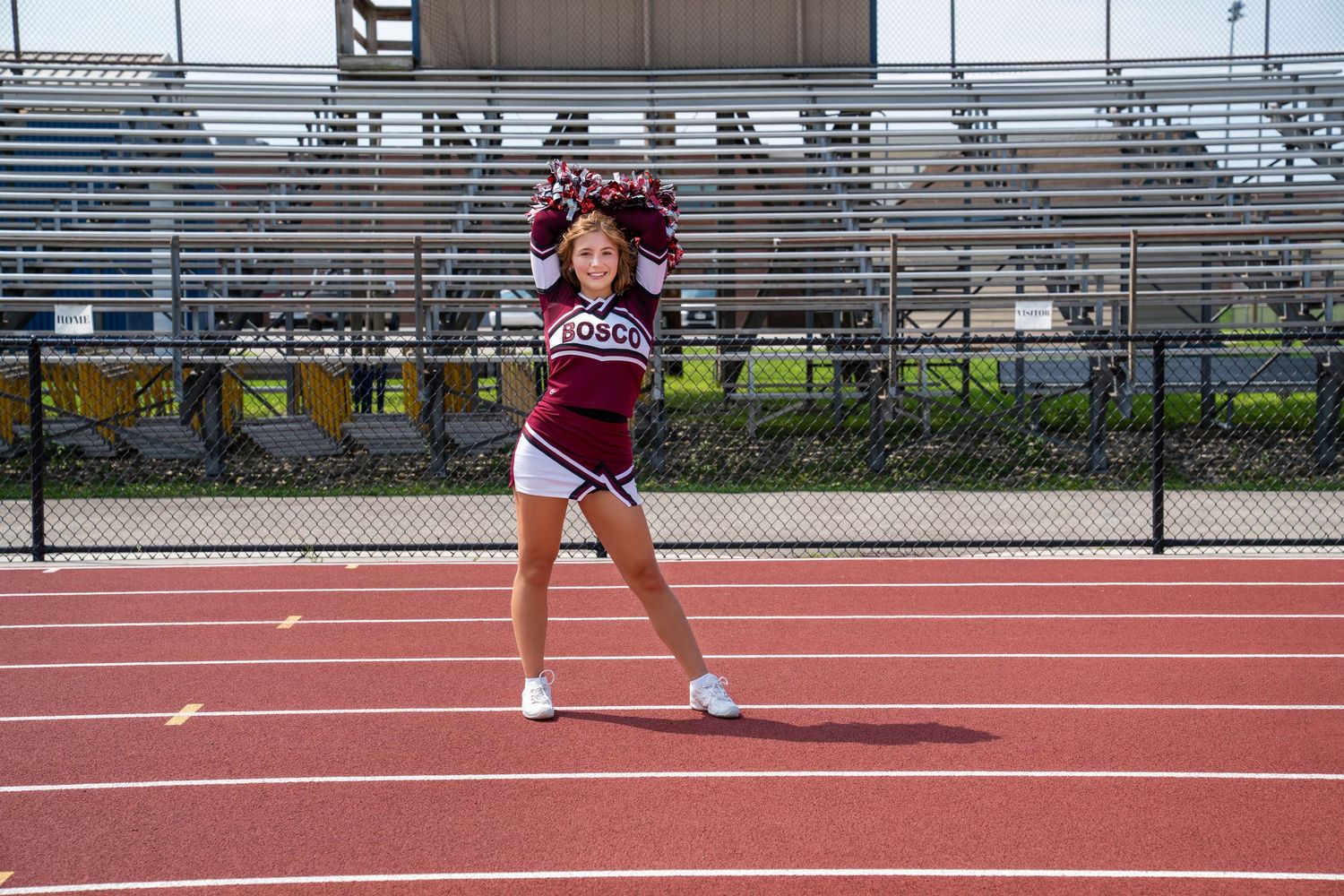 Cheerleader in maroon uniform performs routine on red track with stadium bleachers in background.