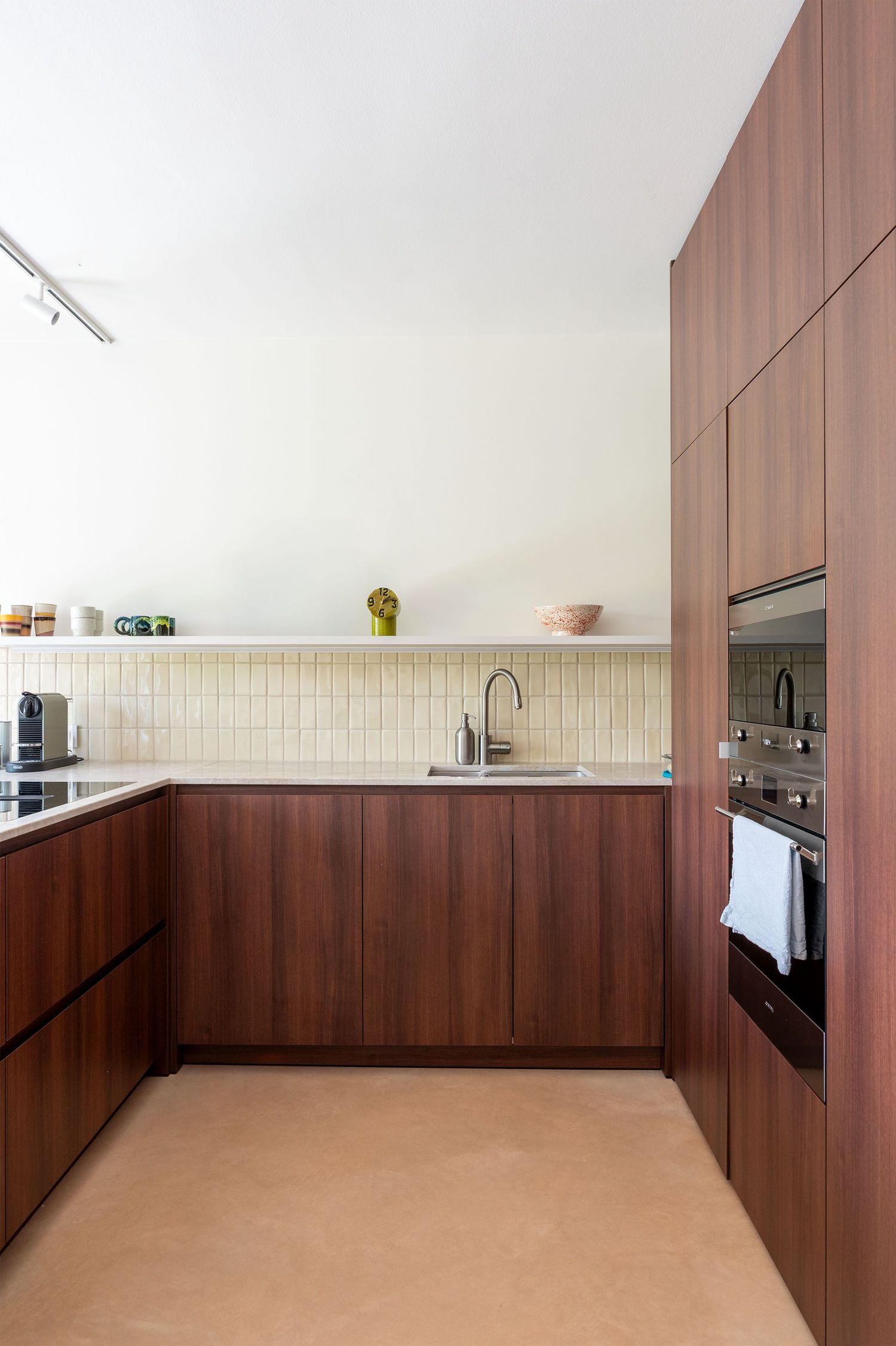 Modern kitchen with walnut cabinets, cream tile backsplash, built-in oven, and cork flooring in a minimalist design.