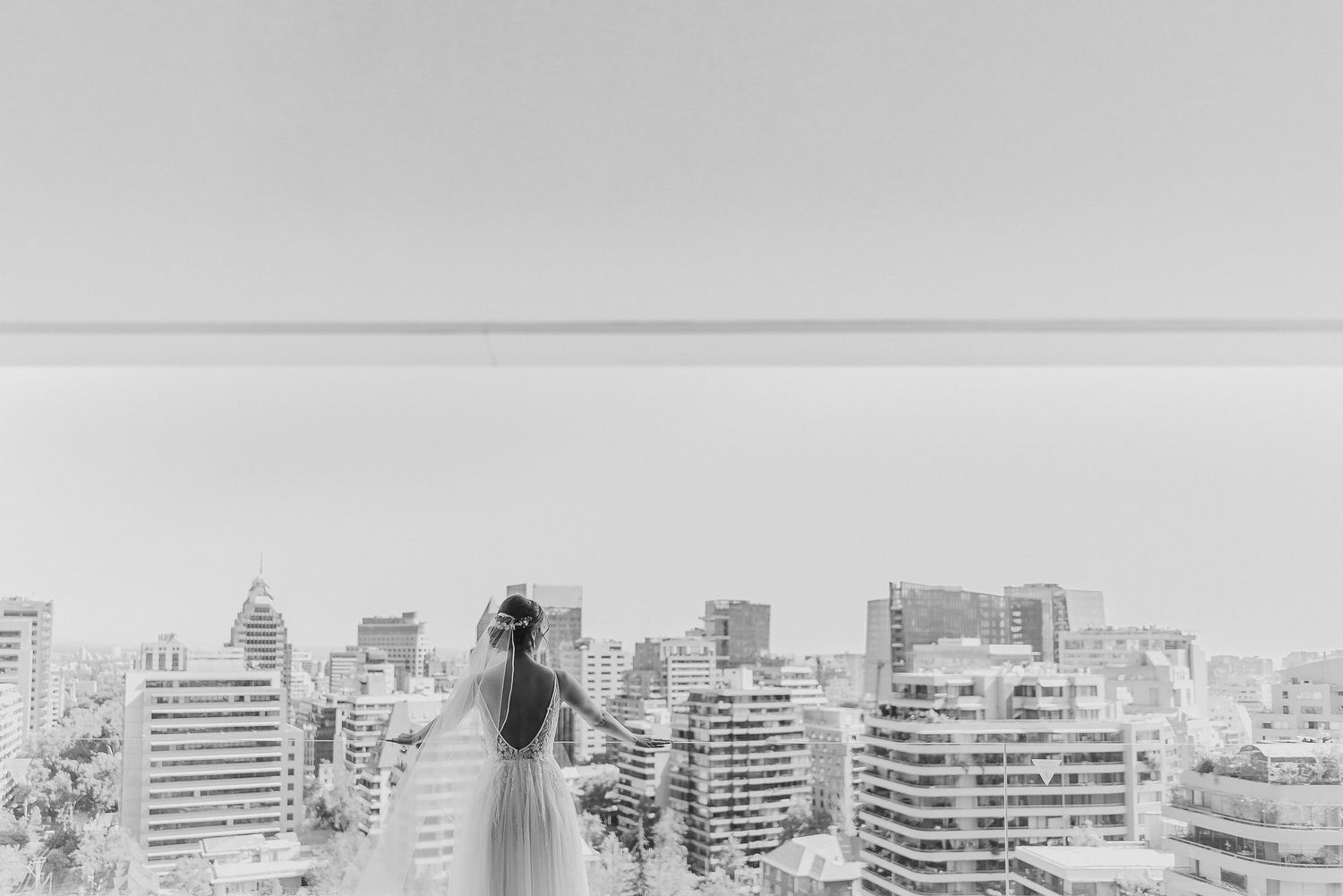 Bride in white wedding gown standing on rooftop overlooking a city skyline in black and white.