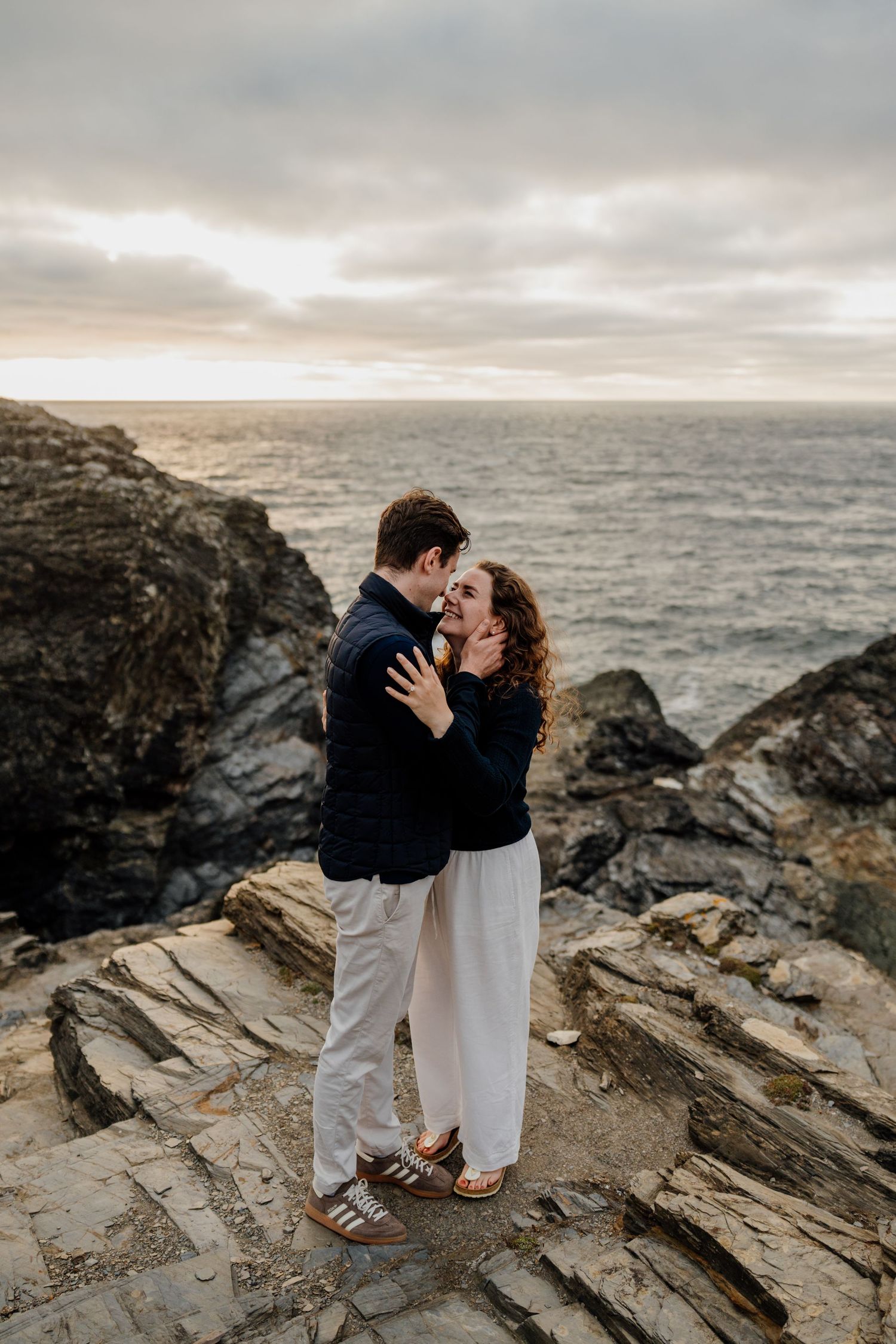 Couple embracing on rocky coastal cliffs at sunset, with dramatic ocean waves and cloudy sky in the background.