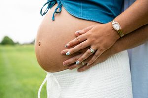 Close-up of a pregnant woman cradling her bare baby bump, wearing a white skirt and gold watch outdoors.