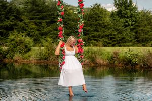 Woman in white dress standing in water holding floral swing with red roses, surrounded by lush green trees.