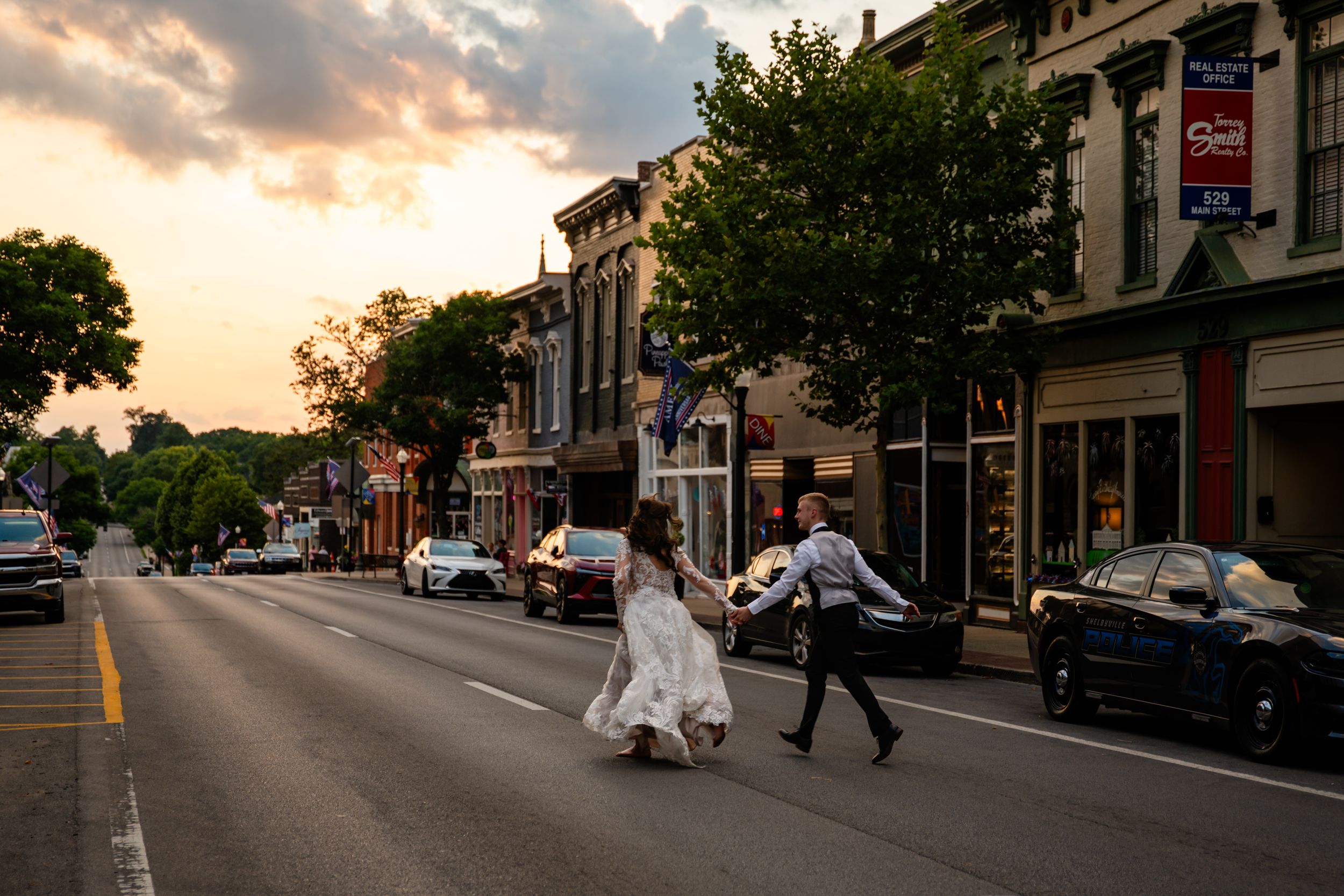 Bride and groom cross a small-town street at sunset, her white gown flowing, golden light illuminating historic storefronts.
