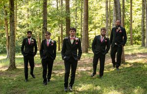 Five groomsmen in black suits with pink boutonnieres pose outdoors among tall trees on a sunny wedding day.