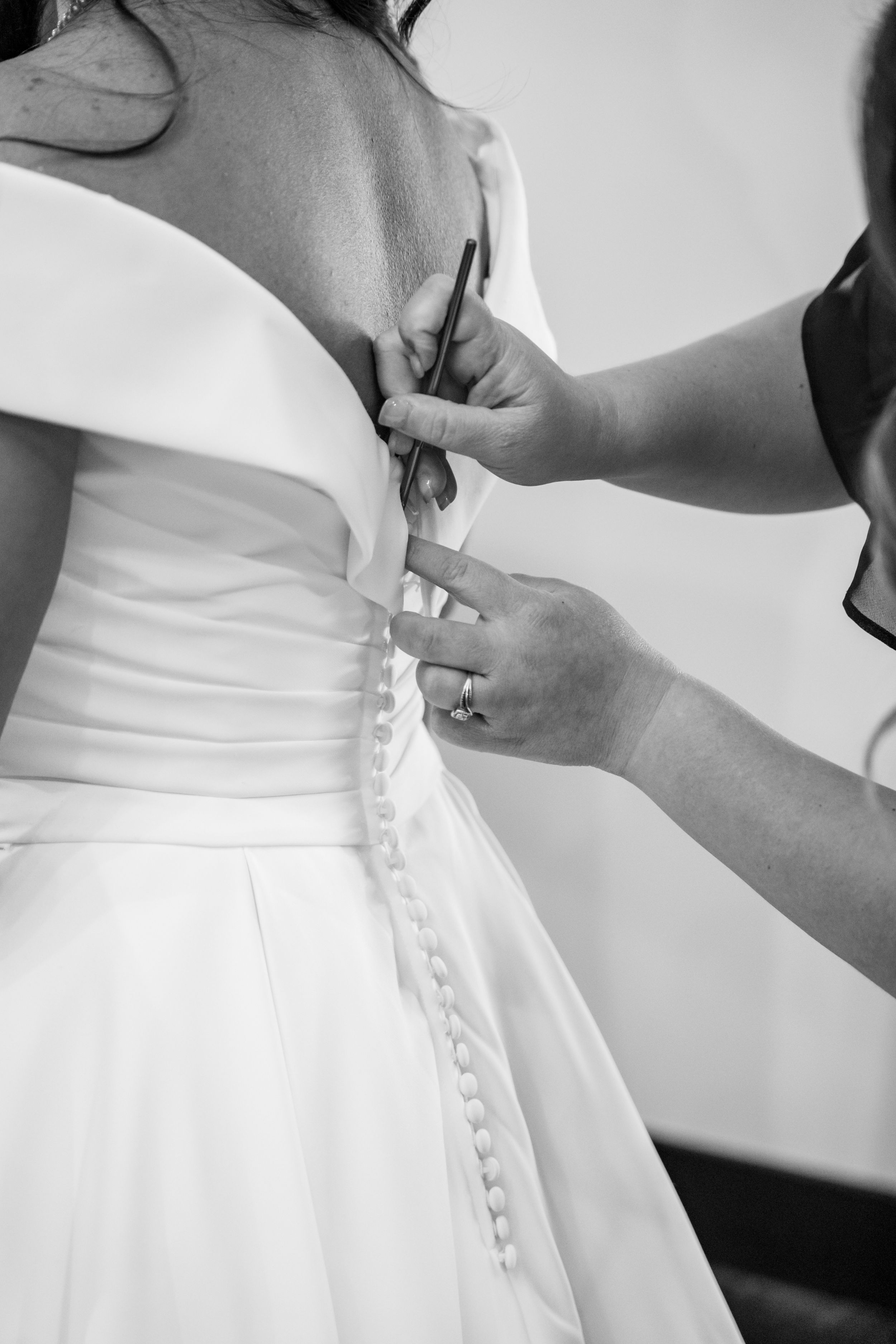 Black and white photo of a bridesmaid buttoning up the back of a wedding dress with buttons.