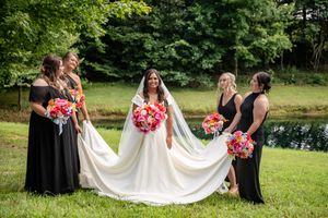 Bride in white gown with bridesmaids in black dresses holding vibrant pink and orange bouquets outdoors near a pond.