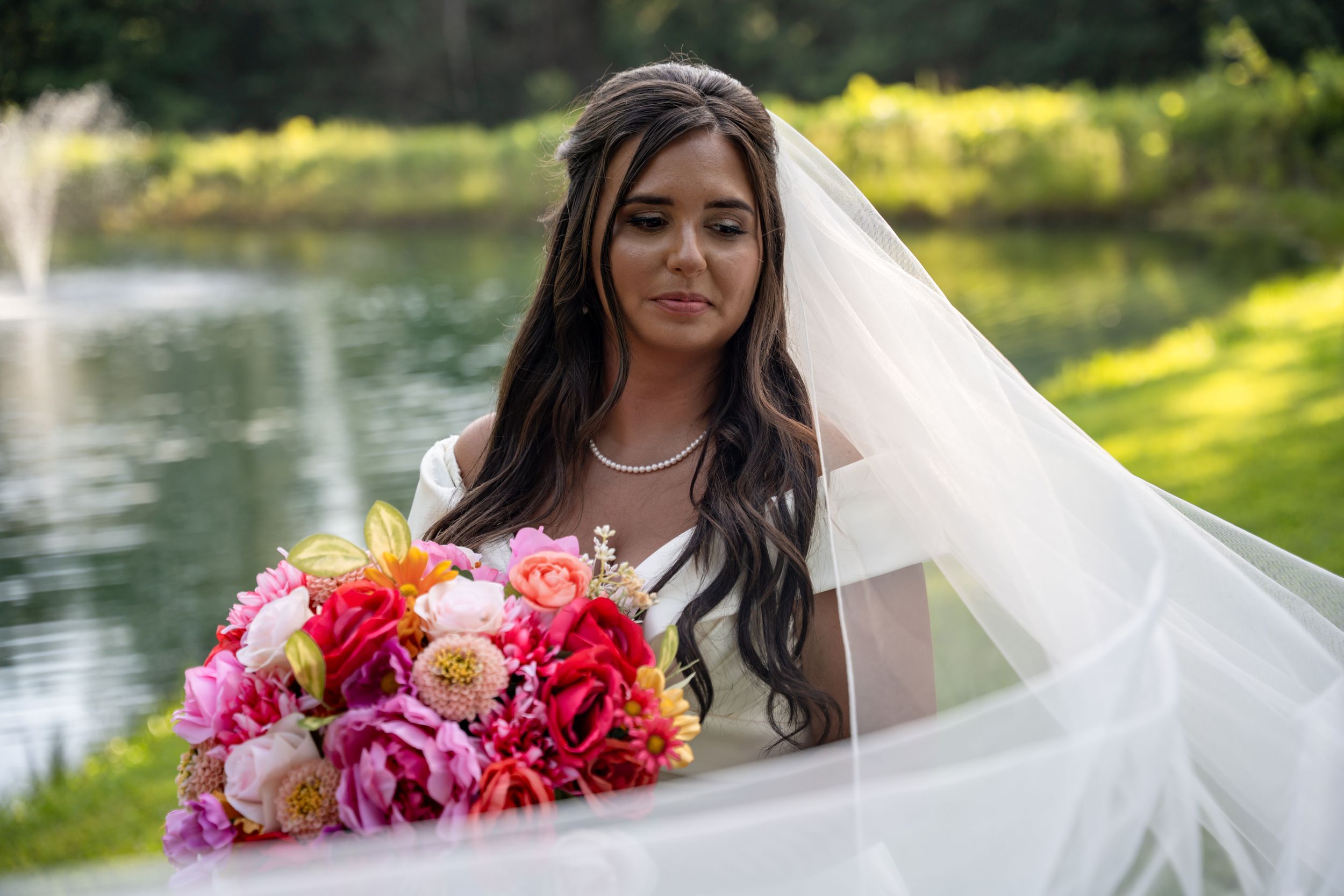 Bride in white gown and veil holds vibrant pink and red bouquet near a serene garden pond.