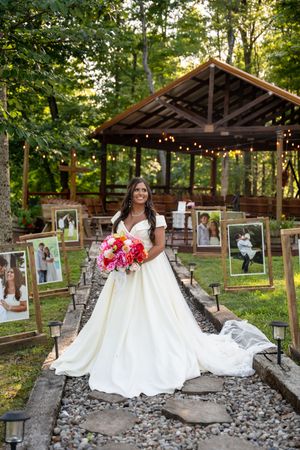 Bride in white ballgown holds colorful bouquet on stone path lined with photos, rustic gazebo with string lights in wooded setting.
