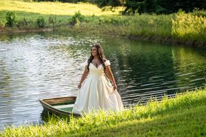 Bride in elegant white gown standing in a wooden boat by a serene pond surrounded by lush green trees.