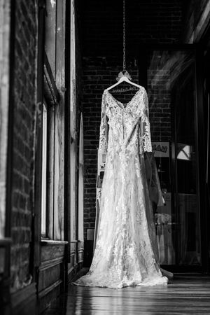 Black and white photo of a lace wedding dress with long sleeves hanging on a personalized hanger in a rustic brick venue.