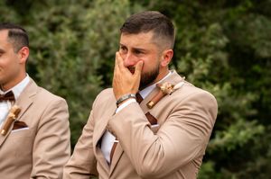Emotional groom in beige suit covering mouth with hand during outdoor wedding ceremony, with groomsman in background.