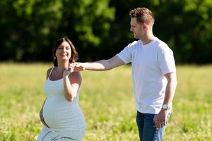 Pregnant woman in white dress holds hands with man in white t-shirt in sunny green field.