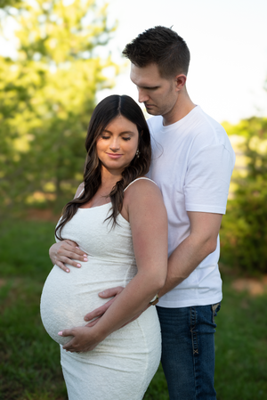 Couple tenderly cradling pregnant belly outdoors in golden light, woman in white dress, man in white tee and jeans.