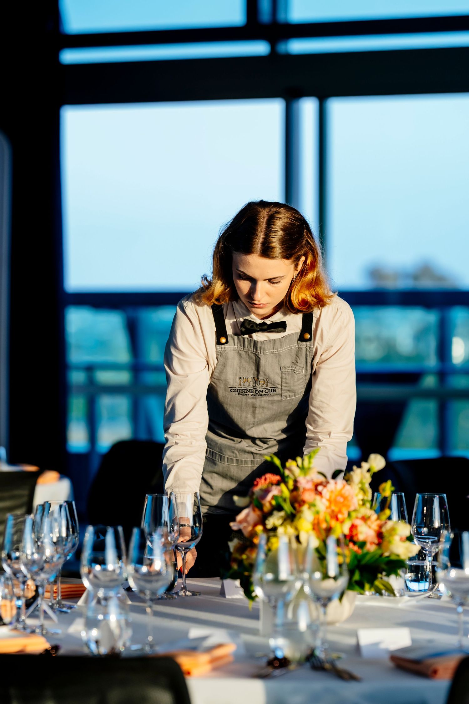 A waitress in an apron arranges wine glasses on an elegantly set table with floral centerpieces in a modern venue.
