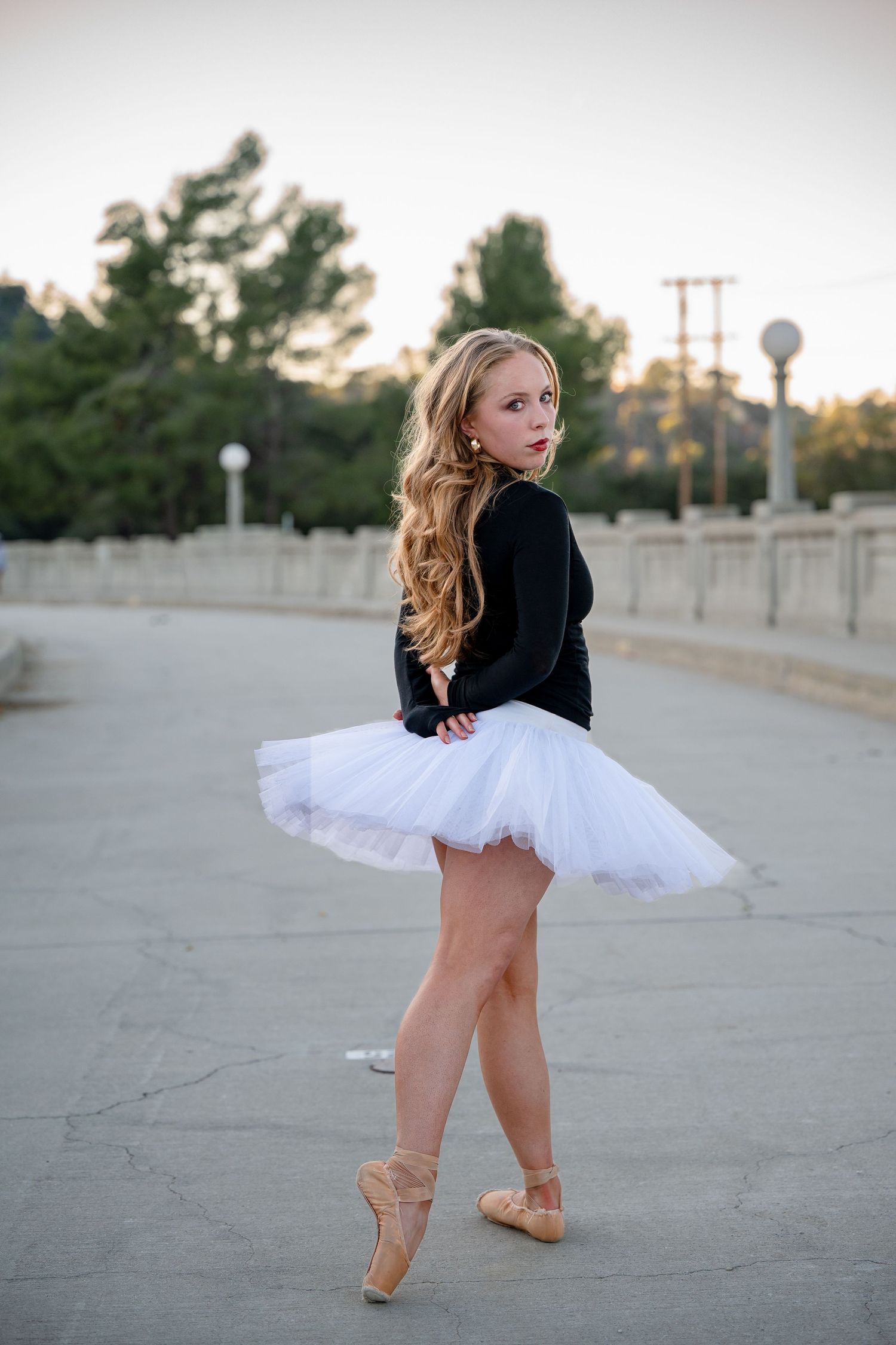Young ballerina in white tutu and pointe shoes poses gracefully outdoors on pavement at sunset.