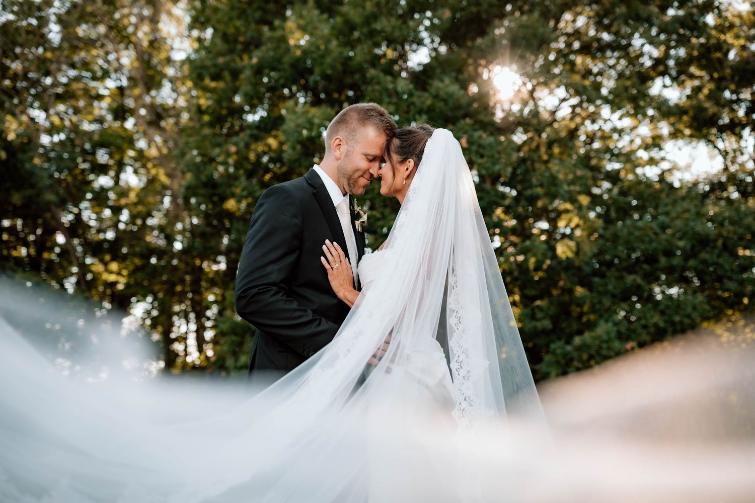 sun peaking through the trees as a bride and groom rest foreheads together with a beautiful veil in the foreground