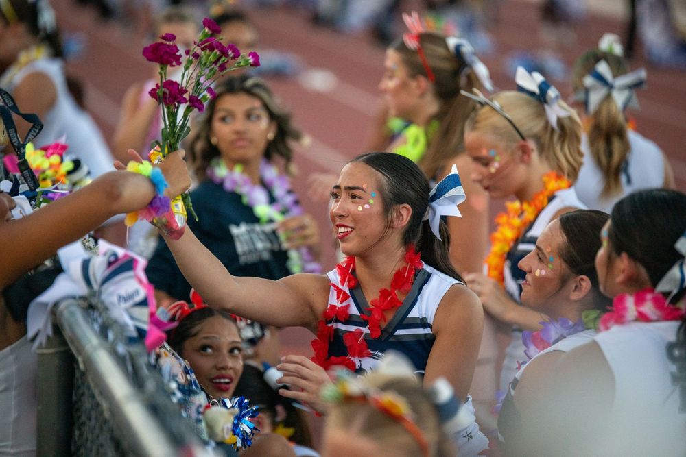 Cheerleaders in uniform celebrate on the sidelines, one smiling girl reaches out to accept colorful flowers from the crowd.