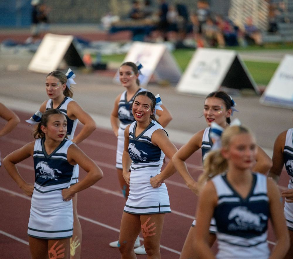 High school cheerleaders in navy and white uniforms perform on a track during a football game.