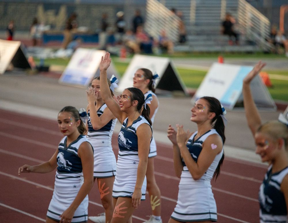 High school cheerleaders in white and navy uniforms perform and wave to the crowd on a red track at a football game.