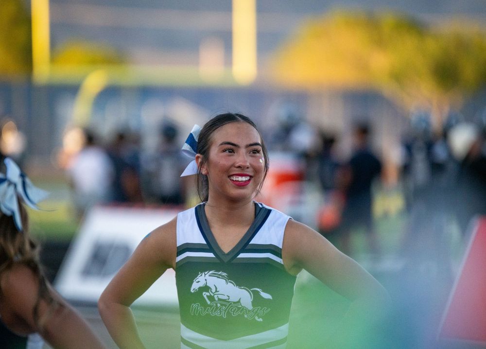 Cheerleader in green and white uniform smiling during a football game on a sunny evening.