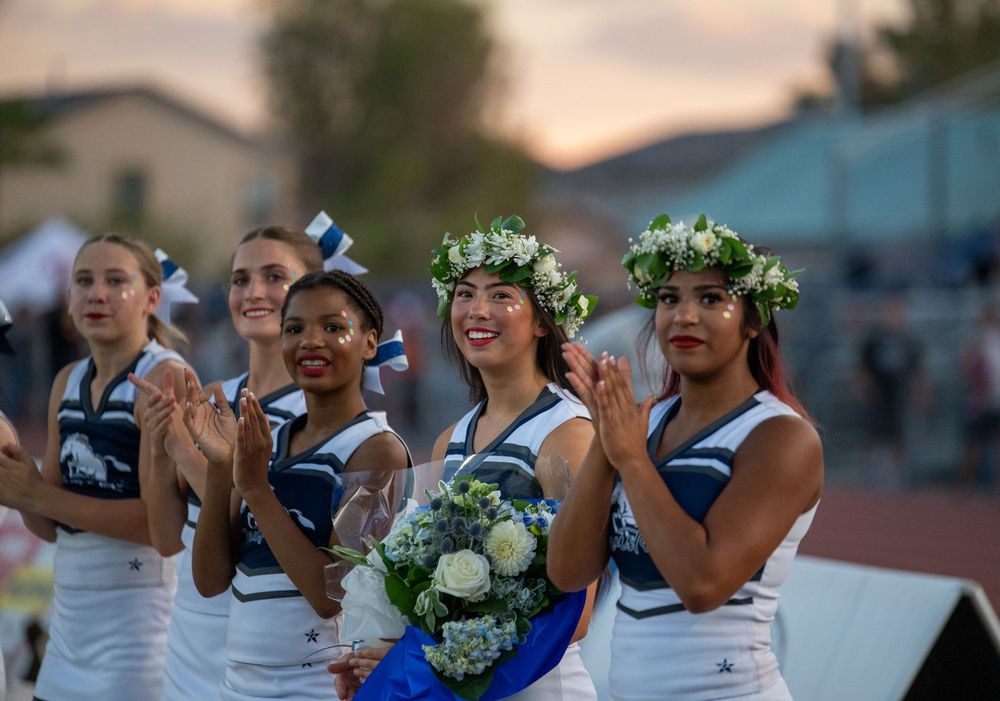 Cheerleaders in blue and white uniforms celebrate at a homecoming event, with one wearing a floral crown and holding a bouquet.