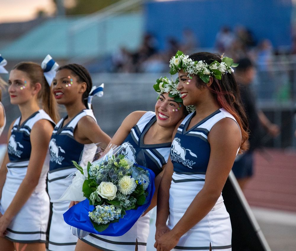 Cheerleader receives floral bouquet and flower crown during senior night ceremony at football game.