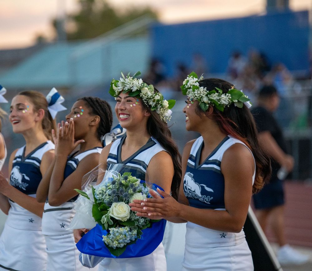 Smiling cheerleader wearing a floral crown holds blue and white bouquet with teammates at a high school football game.