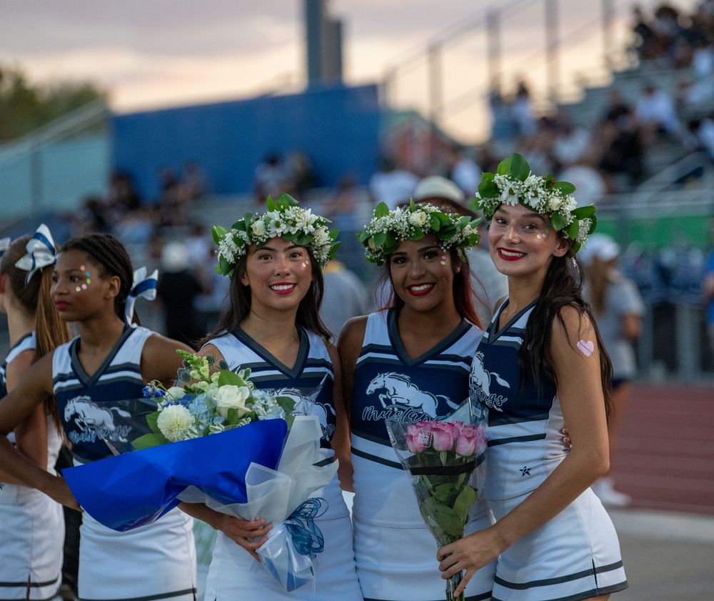 Four cheerleaders in blue and white uniforms wearing floral crowns hold bouquets at a homecoming football game.