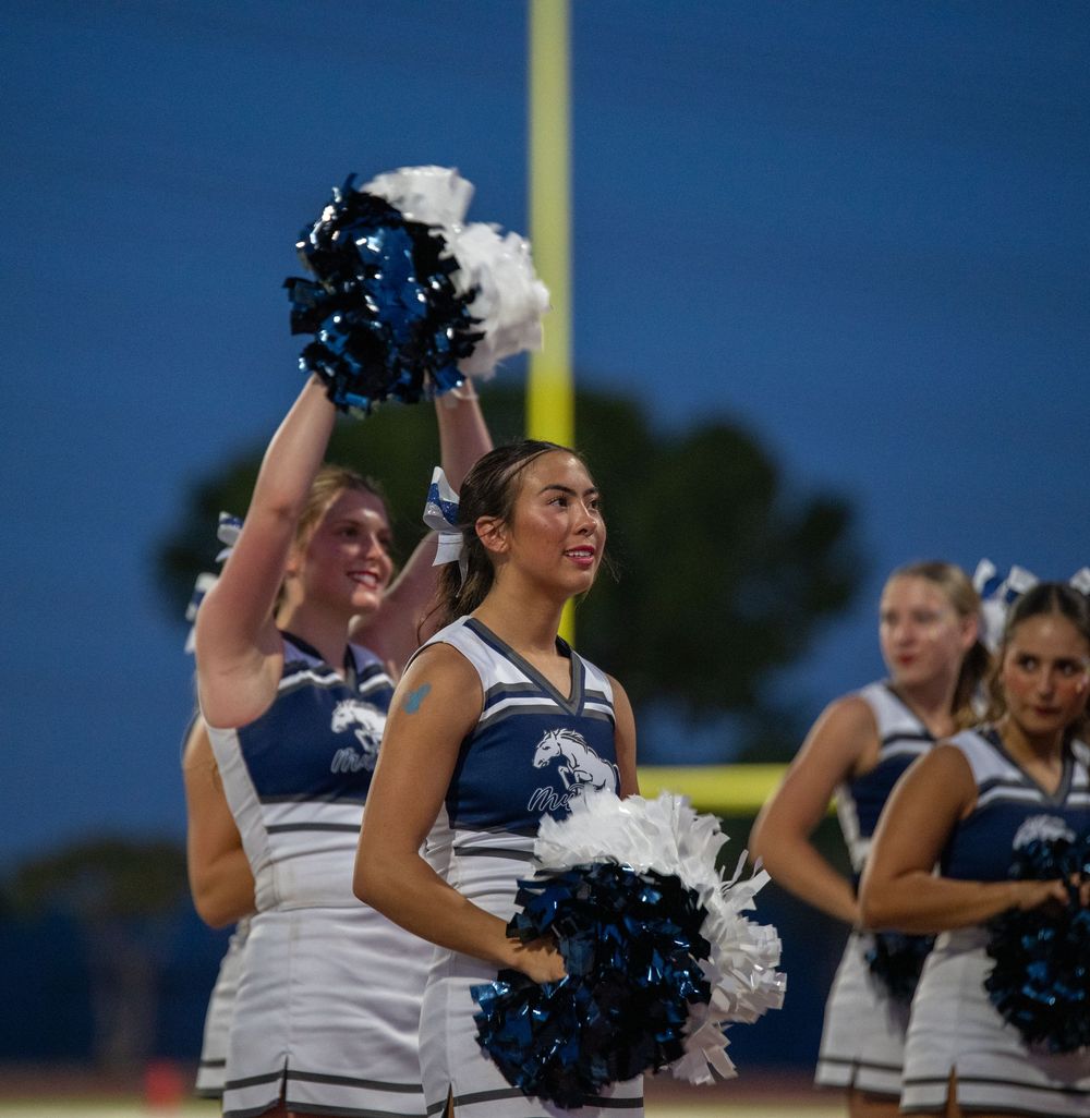 High school cheerleaders in blue and white uniforms wave blue and white pom-poms on the sideline of a football field.