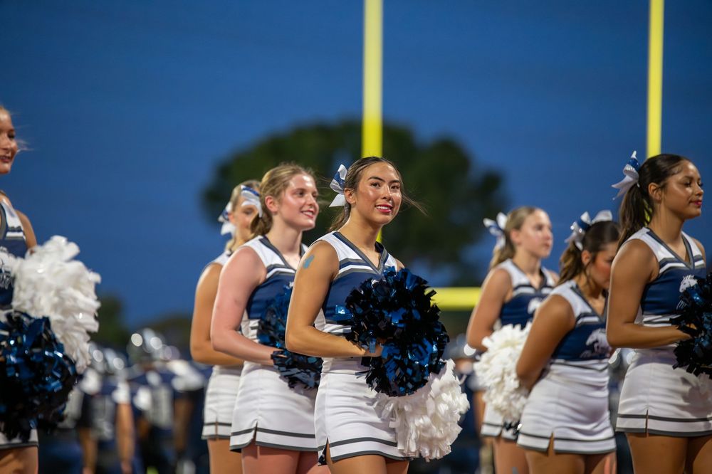 Cheerleaders in white uniforms holding blue and white pom-poms on a football field at night.